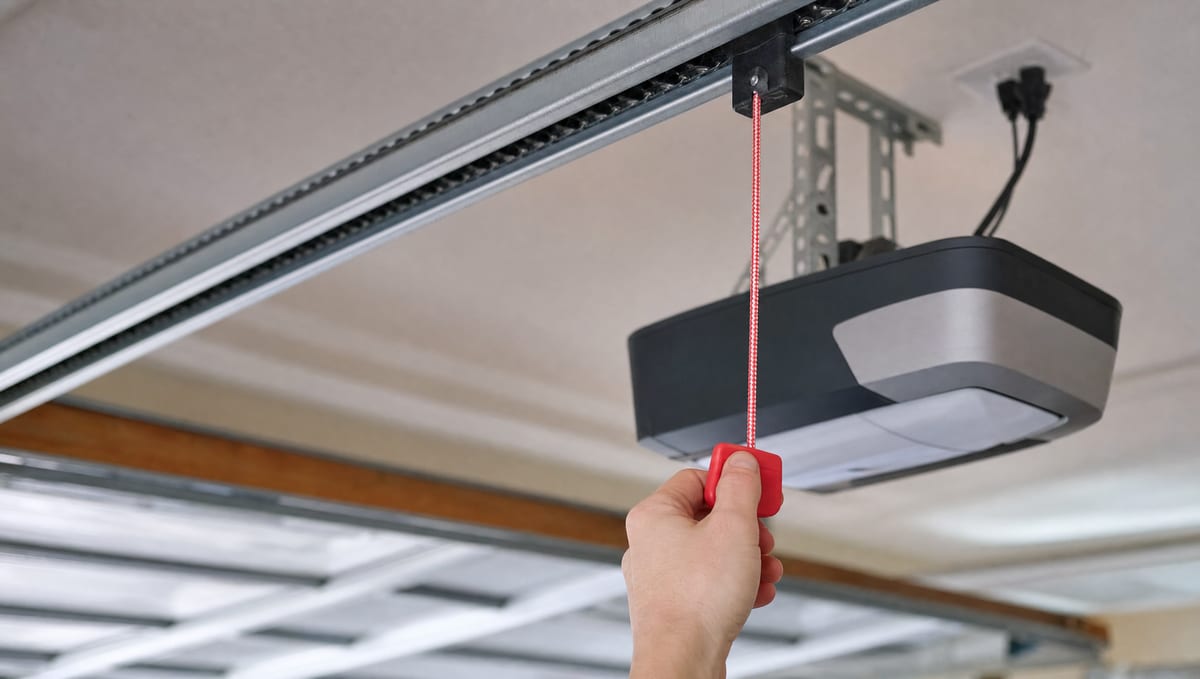 A hand pulling the red manual release cord on a garage door opener rail to disconnect the door